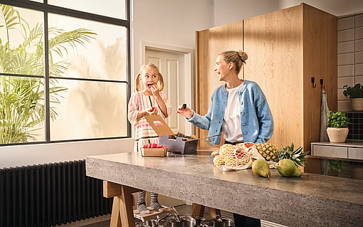 A woman and a child standing in a kitchen, laughing. On the kitchen counter, Mondi-branded corrugated packaging containing fresh fruit.